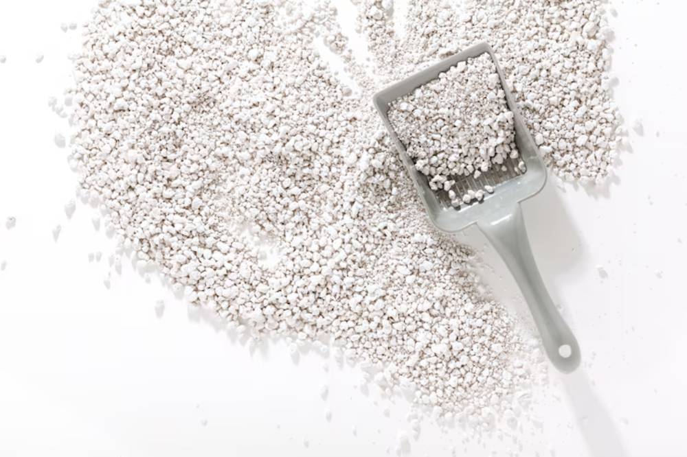 A gray scoop filled with white granular material, surrounded by scattered granules on a white surface, possibly perlite or cat litter.
