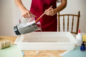 A person in an apron pours blended gray material from a blender into a water-filled tray, making cat litter.