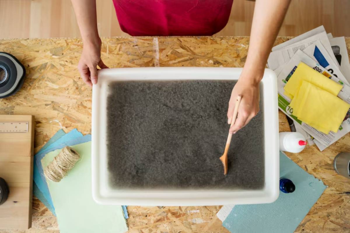 A person stirs dark water in a white tub on a wooden table, surrounded by crafting materials and tools.
