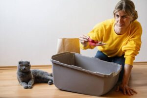A person in a yellow sweater is scooping cat litter into a gray bin with a brown bag nearby, while a gray cat watches curiously.